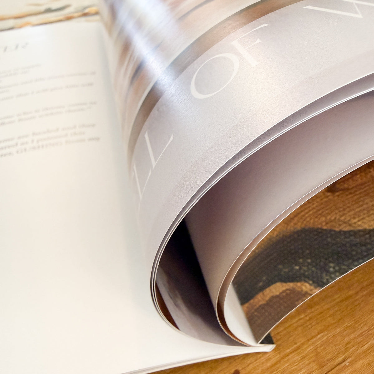 An open art coffee table book lying on a wooden surface, with the right page curled upward to reveal printed pages and a large blurred gray rectangle obscuring part of the content.
