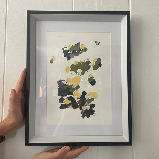 Framed abstract artwork with black, yellow, and green colors on a white wall.