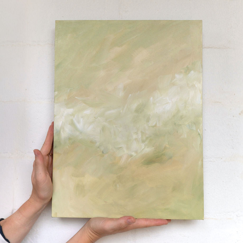 An abstract canvas painting with soft, textured brushstrokes in pale sage green, cream, and beige, held up by two hands against a white brick wall.
